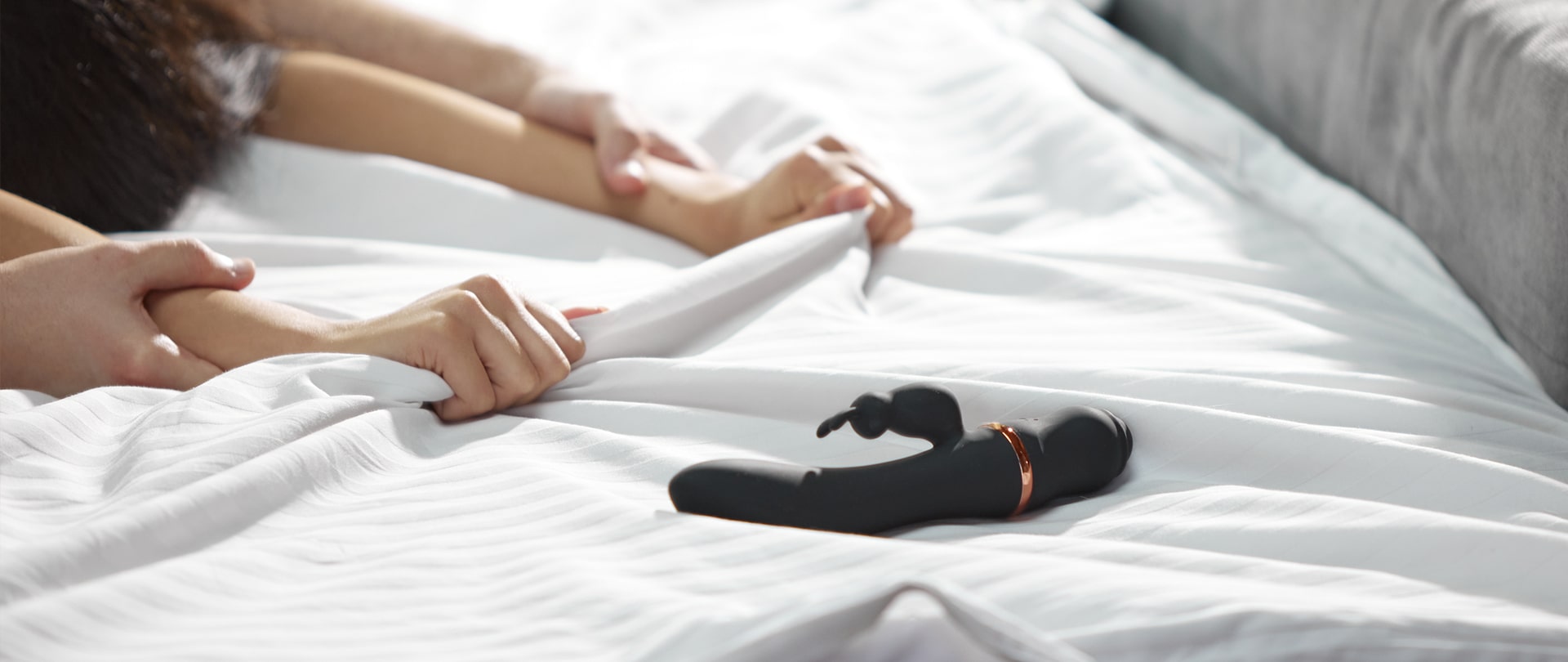 Amina rabbit vibrator i en seng med arme i baggrunden fra et kærestepar der holder ved hinanden
